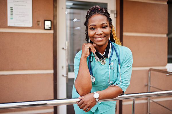 African American woman doctor smiling and leaning over a railing outside of a hospital building