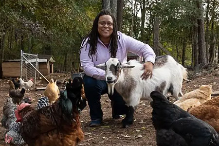 Kim kneeling down with her hand on a goat with chickens scattered walking through her farm