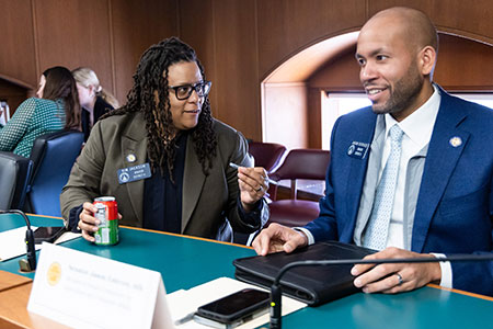 Kim with a soda in her hand looking to her left talking to Senator Jason Esteves who is smiling and listening in a committee meeting