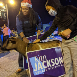 Kim wearing a santa hat leaning over petting a goat next to her yard sign